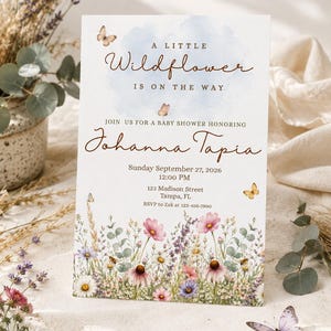 Puede incluir: Una tarjeta de invitación rectangular para baby shower con un fondo de nube azul en acuarela, texto marrón e ilustraciones de mariposas y flores silvestres. El texto dice: A LITTLE Wildflower IS ON THE WAY JOIN US FOR A BABY SHOWER HONORING Johanna Tapia.