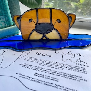 Pode incluir: Uma estatueta de lontra em vitral com cabeça amarelo-castanho e detalhes em preto, repousando sobre uma onda de vidro azul. O design inclui um padrão detalhado para criar a lontra. O texto "3D Otter" é visível.