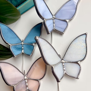 May include: Four stained glass butterfly ornaments with metal solder lines. The butterflies feature translucent glass in shades of light blue, pale lavender, soft pink, and iridescent white, each attached to a thin metal stake for display.