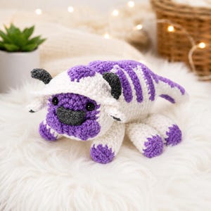 May include: A handmade crochet plush toy featuring a creature with purple and white yarn, dark gray horns, and a dark gray nose. The plush has a rounded head, a long tail, and multiple legs, crafted with visible yarn stitches in a soft texture.
