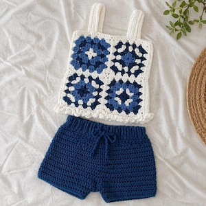 Puede incluir: Conjunto de dos piezas de ganchillo hecho a mano en blanco y azul marino. La parte superior presenta un patrón cuadrado con diseños florales azules, tirantes blancos y un dobladillo con volantes. Los pantalones cortos a juego son de color azul marino liso con cintura con cordón.