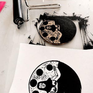 Op de afbeelding: Een zwart-wit linoleumsnede van een figuur in een cirkel, met een roller en inkt op een helder oppervlak. De prent toont een gestileerde menselijke vorm met een donker silhouet en lichte details. De afbeelding suggereert grafisch drukken.