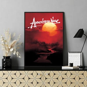 Op de afbeelding: Een ingelijste poster met de filmtitel "Apocalypse Now" in witte letters tegen een rode en oranje zonsondergang. Het kunstwerk toont een rivierlandschap met silhouetbomen en een heldere zon.
