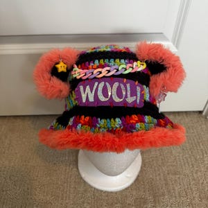May include: A crocheted bucket hat featuring horizontal stripes of black and multicoloured yarn, trimmed with bright orange faux fur. It includes a purple patch with the word WOOL in glittery letters, a pastel plastic chain, and two fuzzy orange ears with star charms.
