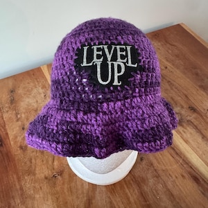 May include: A crocheted bucket hat in variegated shades of purple, featuring a black patch on the front with the text "LEVEL UP" in silver lettering. The hat has a wide, flexible brim and a textured, hand-stitched appearance throughout the design.