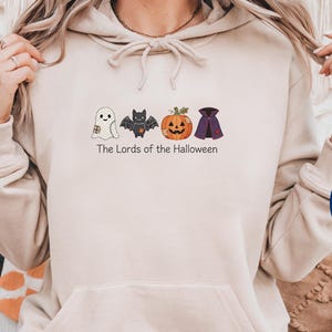 May include: A beige hooded sweatshirt featuring a graphic print of a ghost, a bat, a jack-o'-lantern, and a cape. Below the icons, the text reads "The Lords of the Halloween". The hoodie has long sleeves, a front kangaroo pocket, and a drawstring hood.