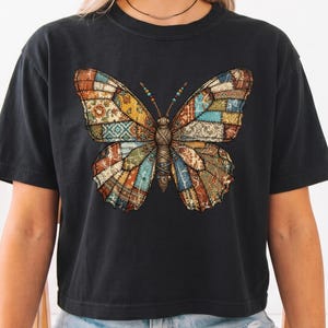 May include: A black short-sleeved t-shirt featuring a large, central graphic of a butterfly. The butterfly wings are composed of a patchwork design with various intricate patterns in shades of brown, blue, yellow, and orange. The shirt has a crew neckline.