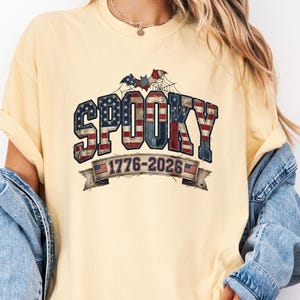 Peut inclure: Un t-shirt à manches courtes jaune beurre avec le mot SPOOKY écrit dans un motif de drapeau américain rouge, blanc et bleu. Au-dessus du texte se trouve une chauve-souris et une toile d'araignée. Une bannière affiche les années 1776-2026.