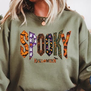 May include: A military green long-sleeve sweatshirt featuring the word SPOOKY in large, multi-patterned, stitched-style letters with Halloween motifs like ghosts, pumpkins, and spiderwebs, and the word HALLOWEEN printed in orange below.