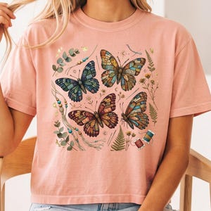 May include: A peach-coloured short-sleeve t-shirt featuring a graphic print of four butterflies with intricate stained-glass style patterns. The design includes botanical elements like fern leaves, eucalyptus sprigs, small flowers, sewing needles, and spools of thread.