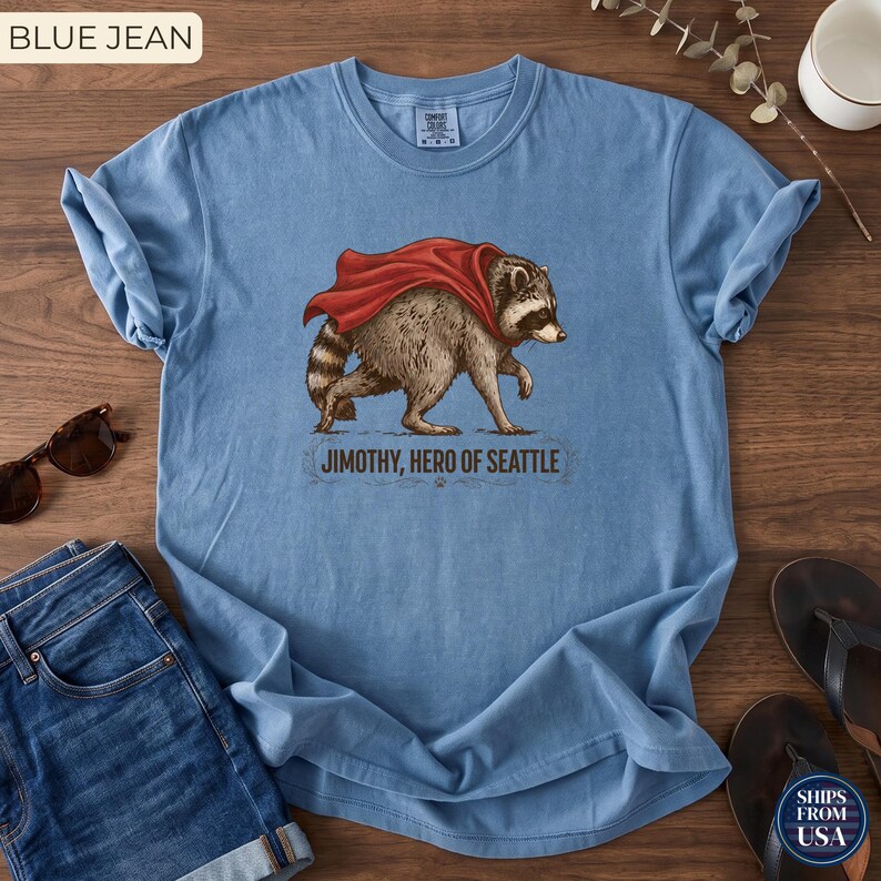 May include: A blue short-sleeve t-shirt featuring a graphic of a raccoon wearing a red cape. Below the raccoon, the text reads "JIMOTHY, HERO OF SEATTLE". The shirt has a crew neckline and a relaxed fit, showcasing a detailed illustration of the animal.
