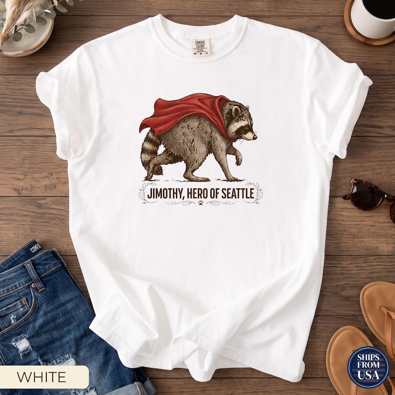 May include: A white short-sleeve t-shirt featuring a graphic of a raccoon wearing a red cape. Below the raccoon, the text reads JIMOTHY, HERO OF SEATTLE, accompanied by a small paw print icon and decorative flourishes on either side of the text.