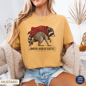 May include: A mustard yellow short-sleeve t-shirt featuring a graphic illustration of a raccoon wearing a red cape. Below the raccoon, the text reads "JIMOTHY, HERO OF SEATTLE" in a dark brown font, accented by decorative flourishes and a small paw print.