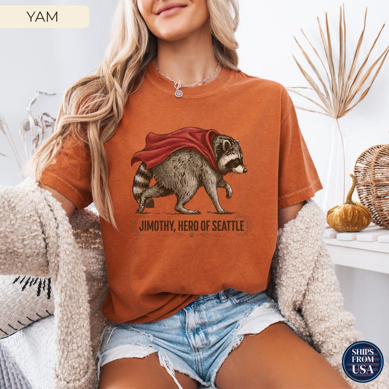 May include: A burnt orange short-sleeve t-shirt featuring a graphic illustration of a raccoon wearing a red cape. Below the raccoon, the text reads: JIMOTHY, HERO OF SEATTLE. The shirt has a crew neckline and a relaxed, casual fit.