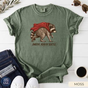 May include: A moss green crew neck t-shirt featuring a graphic illustration of a raccoon wearing a red cape. Below the raccoon, the text reads JIMOTHY, HERO OF SEATTLE in dark brown lettering. The shirt has a classic fit with short sleeves.