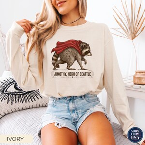 May include: An ivory long-sleeve sweatshirt featuring a graphic illustration of a raccoon wearing a red cape. Below the raccoon, the text reads JIMOTHY, HERO OF SEATTLE, accompanied by decorative flourishes and a small paw print icon.