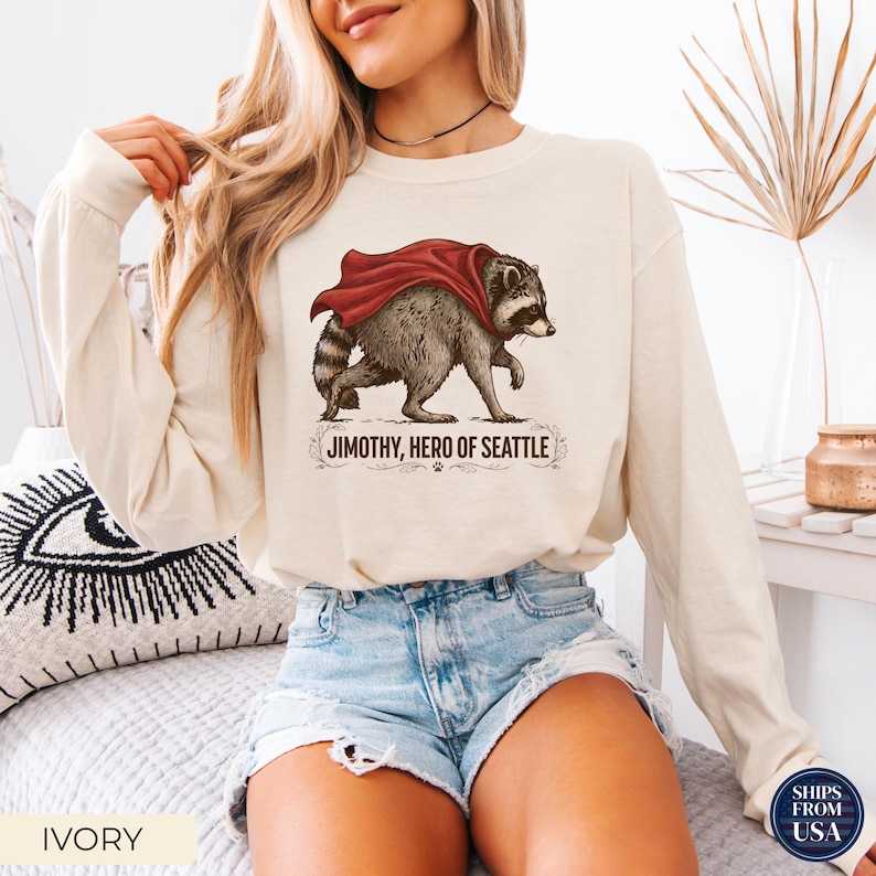 May include: An ivory long-sleeve sweatshirt featuring a graphic illustration of a raccoon wearing a red cape. Below the raccoon, the text reads JIMOTHY, HERO OF SEATTLE, accompanied by decorative flourishes and a small paw print icon.