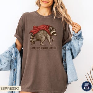 May include: An espresso-colored crew neck t-shirt featuring a graphic illustration of a raccoon wearing a red cape. Below the raccoon, the text reads JIMOTHY, HERO OF SEATTLE in a dark brown font. The shirt has a relaxed fit and short sleeves.