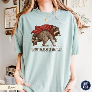 May include: A light sage green t-shirt featuring a graphic of a raccoon wearing a red cape. Below the raccoon, the text reads JIMOTHY, HERO OF SEATTLE, accompanied by a small paw print and decorative flourishes. The shirt has a crew neckline.