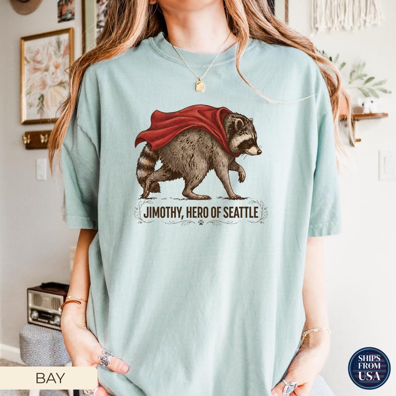 May include: A light sage green t-shirt featuring a graphic of a raccoon wearing a red cape. Below the raccoon, the text reads JIMOTHY, HERO OF SEATTLE, accompanied by a small paw print and decorative flourishes. The shirt has a crew neckline.