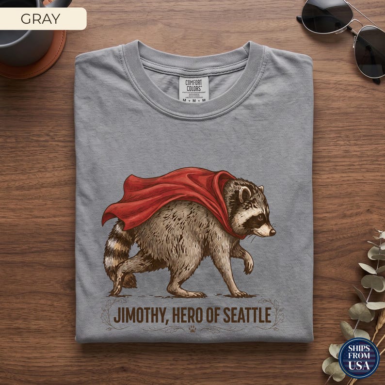 May include: A gray crew neck t-shirt featuring a graphic illustration of a raccoon wearing a flowing red cape. Below the raccoon, the text reads JIMOTHY, HERO OF SEATTLE. The shirt has a Comfort Colors brand label visible at the inner neckline.