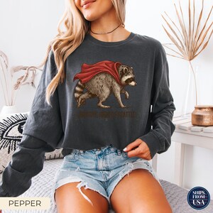 May include: A charcoal gray long-sleeve sweatshirt featuring a graphic of a raccoon wearing a red cape. Below the raccoon, the text reads "TIMOTHY, HERO OF SEATTLE". The sweatshirt has a crew neckline and a relaxed fit with long sleeves.