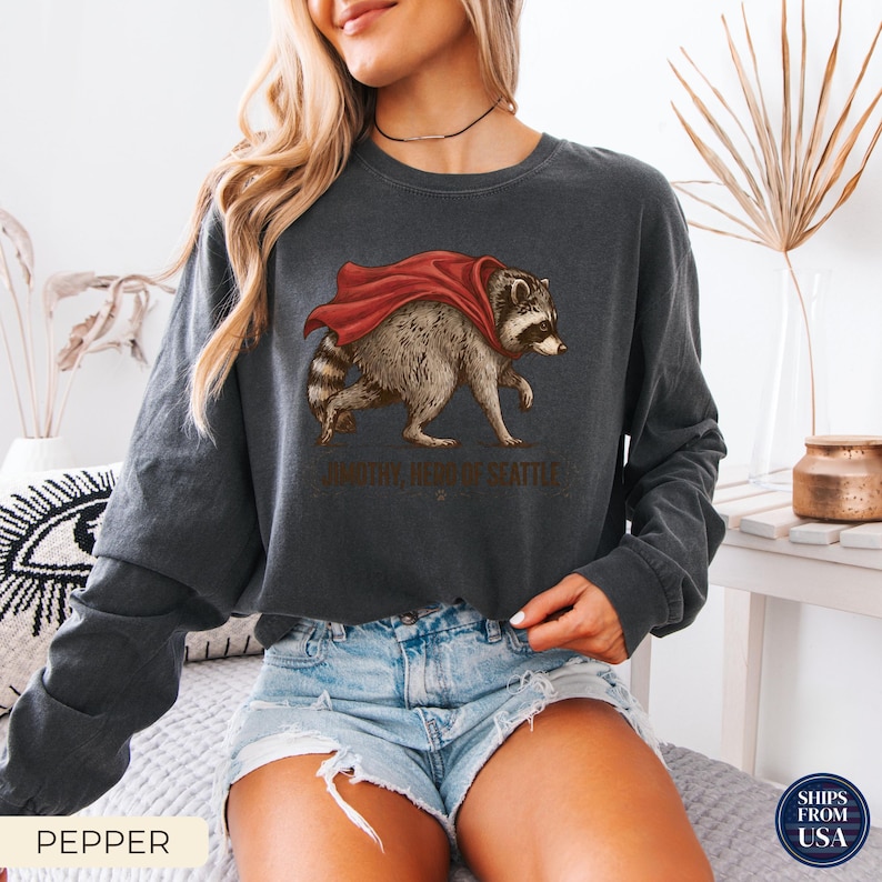 May include: A charcoal gray long-sleeve sweatshirt featuring a graphic of a raccoon wearing a red cape. Below the raccoon, the text reads "TIMOTHY, HERO OF SEATTLE". The sweatshirt has a crew neckline and a relaxed fit with long sleeves.
