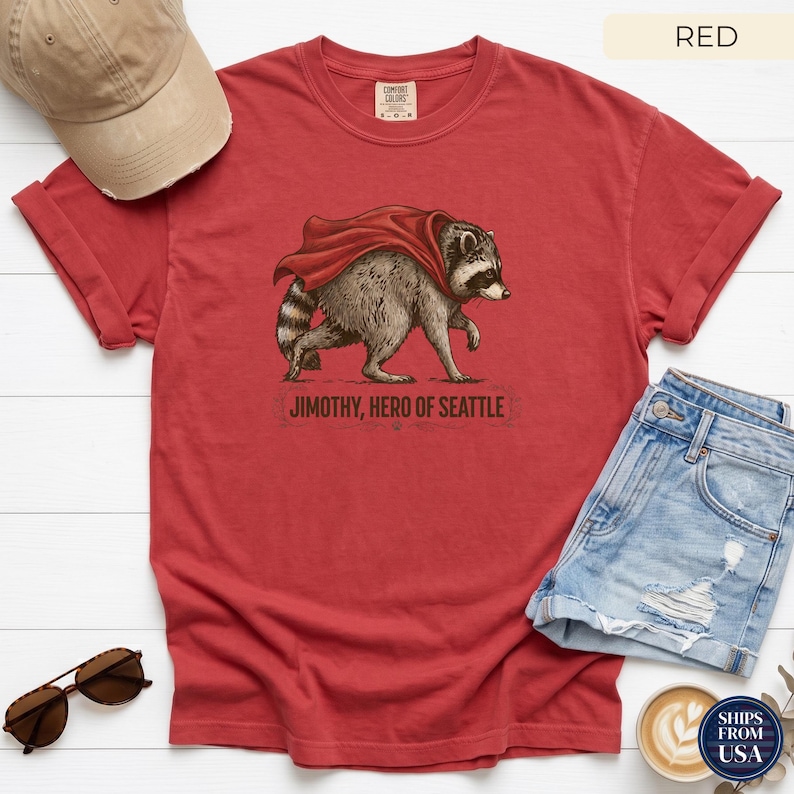 May include: A red short-sleeve t-shirt featuring a graphic of a raccoon wearing a red cape. Below the raccoon, the text reads "JIMOTHY, HERO OF SEATTLE". The shirt has a crew neckline and a relaxed fit, displayed with a tan baseball cap and denim shorts.