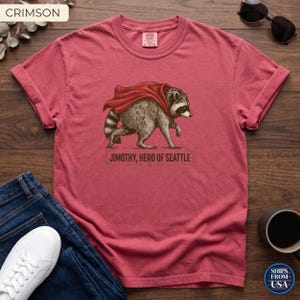 May include: A crimson short-sleeve t-shirt featuring a graphic of a raccoon wearing a red cape. Below the raccoon, the text reads JIMOTHY, HERO OF SEATTLE. The shirt has a crew neckline and a label at the inner collar area.