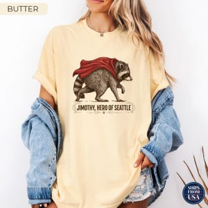 May include: A butter-colored t-shirt featuring a graphic of a raccoon wearing a red cape. Below the raccoon, the text reads "JIMOTHY, HERO OF SEATTLE" in black capital letters, framed by decorative flourishes and a small paw print icon.