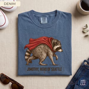 May include: A denim blue crew neck t-shirt featuring a graphic illustration of a raccoon wearing a red cape. Below the raccoon, the text reads "JIMOTHY, HERO OF SEATTLE" in a dark brown font. The shirt has a Comfort Colors brand label at the neck.