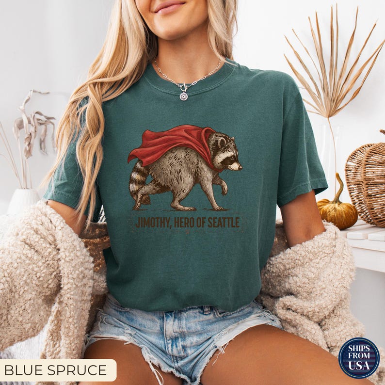 May include: A dark teal short-sleeve t-shirt featuring a graphic of a raccoon wearing a red cape. Below the raccoon, the text reads JIMOTHY, HERO OF SEATTLE. The shirt has a crew neckline and a relaxed fit, showcasing a detailed illustration.