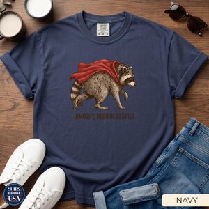 May include: A navy blue short-sleeve t-shirt featuring a graphic of a raccoon wearing a red cape. Below the raccoon, the text reads JIMOTHY, HERO OF SEATTLE. The shirt has a crew neckline and a Comfort Colors brand label on the inside collar.