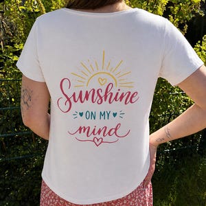 Könnte beinhalten: Cremefarbenes T-Shirt mit dem Schriftzug "Sunshine on my mind" in Rosa und Türkis, mit einer gelben Sonnengrafik. Das Shirt hat einen Rundhalsausschnitt und kurze Ärmel.