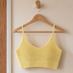 May include: A yellow crochet crop top with a V-neck and thin straps, hanging on a wooden hanger. The top is made of a textured, open-weave fabric. The background is a light beige wall.