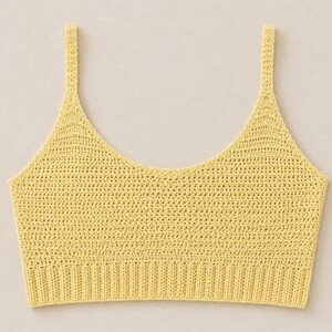May include: A yellow crochet top with a scoop neckline and thin shoulder straps. The top has a ribbed band at the bottom and a delicate, open weave throughout the body. The color is a soft, pastel yellow.