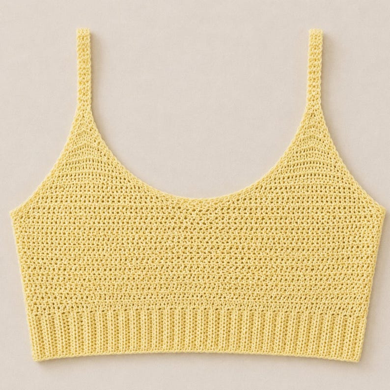May include: A yellow crochet top with a scoop neckline and thin shoulder straps. The top has a ribbed band at the bottom and a delicate, open weave throughout the body. The color is a soft, pastel yellow.