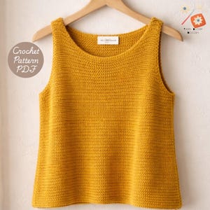 May include: Mustard yellow crochet tank top hanging on a wooden hanger. The sleeveless top has a scoop neckline and a textured stitch pattern. The words "Crochet Pattern PDF" are on the left side of the image. A small tag is visible near the neckline.