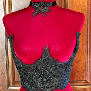 May include: A black lace corset top featuring intricate embroidery, floral patterns, and dark bead embellishments, paired with a matching black velvet choker adorned with a central lace and beaded floral applique on a red mannequin.
