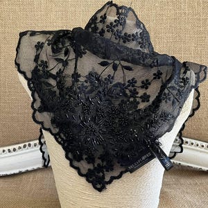 May include: A black lace scarf with floral and beaded detailing. The scarf is displayed on a mannequin head, highlighting its intricate design and scalloped edges. A tag with the brand name is attached.