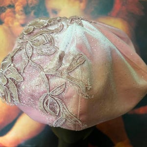May include: A soft pink velvet beret featuring an intricate beige floral lace appliqué on one side. The fabric has a subtle shimmer and is adorned with tiny, scattered sequins that catch the light, creating a delicate and textured appearance.