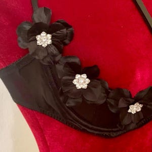 May include: Black bra with floral embellishments. The bra features three large black fabric flowers with sparkling rhinestone centers. The bra is displayed on a red mannequin, showcasing its design and details. The bra has a delicate strap design.