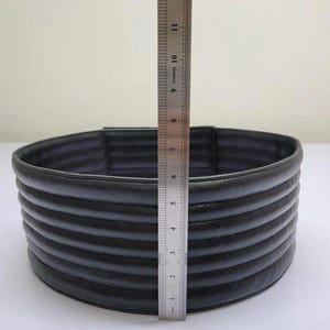 Puede incluir: Un cintur&oacute;n negro, cil&iacute;ndrico y acolchado con canales cosidos horizontalmente. Una regla de acero inoxidable est&aacute; colocada verticalmente contra el frente, mostrando que el cintur&oacute;n tiene una altura de aproximadamente 10 cm desde el borde inferior al superior.