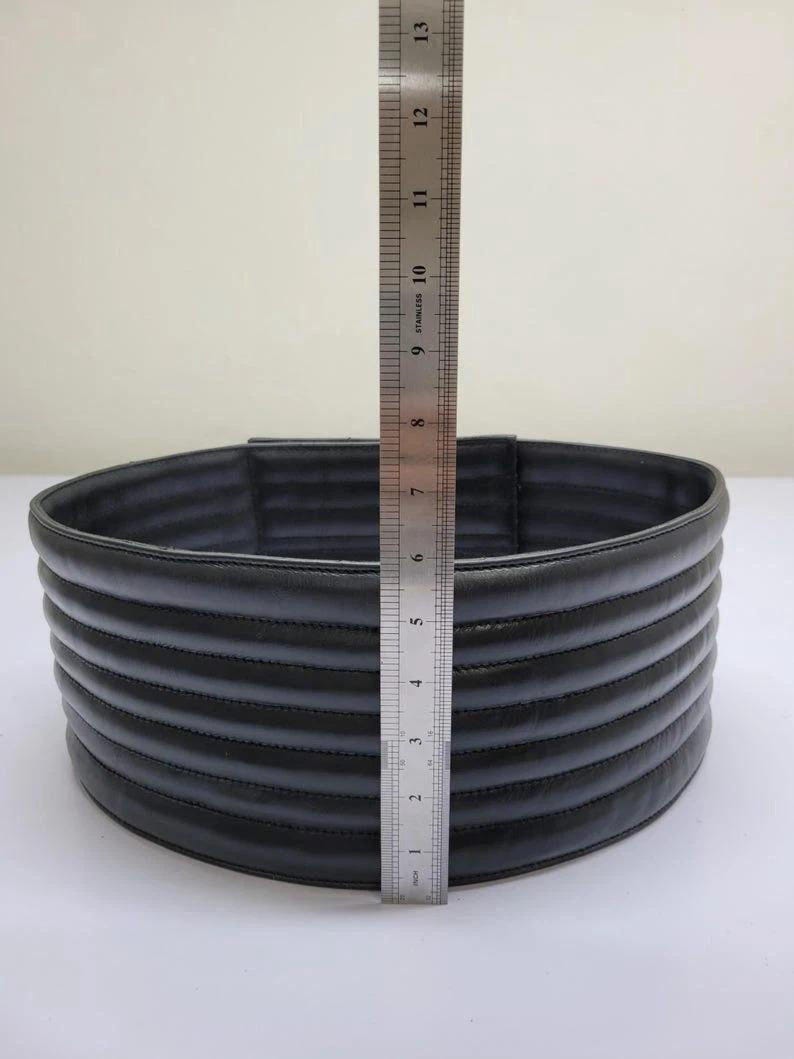 Puede incluir: Un cintur&oacute;n negro, cil&iacute;ndrico y acolchado con canales cosidos horizontalmente. Una regla de acero inoxidable est&aacute; colocada verticalmente contra el frente, mostrando que el cintur&oacute;n tiene una altura de aproximadamente 10 cm desde el borde inferior al superior.