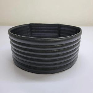 Puede incluir: Un cintur&oacute;n ancho de color negro que presenta una serie de canales horizontales acolchados y acolchados a lo largo de toda su longitud. El cintur&oacute;n tiene un acabado mate liso y una forma cil&iacute;ndrica estructurada con costuras visibles.