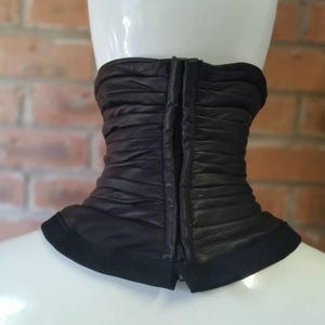 May include: A black leather neck gaiter featuring horizontal ruched detailing and a central vertical zipper closure. The accessory has a smooth, solid black fabric trim along the bottom edge and is displayed on a white mannequin neck form.