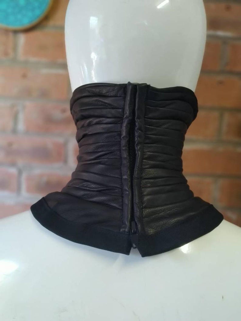 May include: A black leather neck gaiter featuring horizontal ruched detailing and a central vertical zipper closure. The accessory has a smooth, solid black fabric trim along the bottom edge and is displayed on a white mannequin neck form.