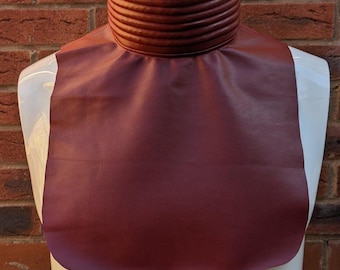 May include: A dark red faux leather dickey featuring a high, ribbed mock neck collar and a smooth, rounded bib front that covers the upper chest area. The accessory is displayed on a white mannequin against a red brick wall background.
