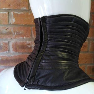 May include: A dark brown leather neck gaiter featuring horizontal pleated detailing and a vertical front zipper closure. The accessory is designed to fit snugly around the neck, providing a structured appearance with a smooth finish.