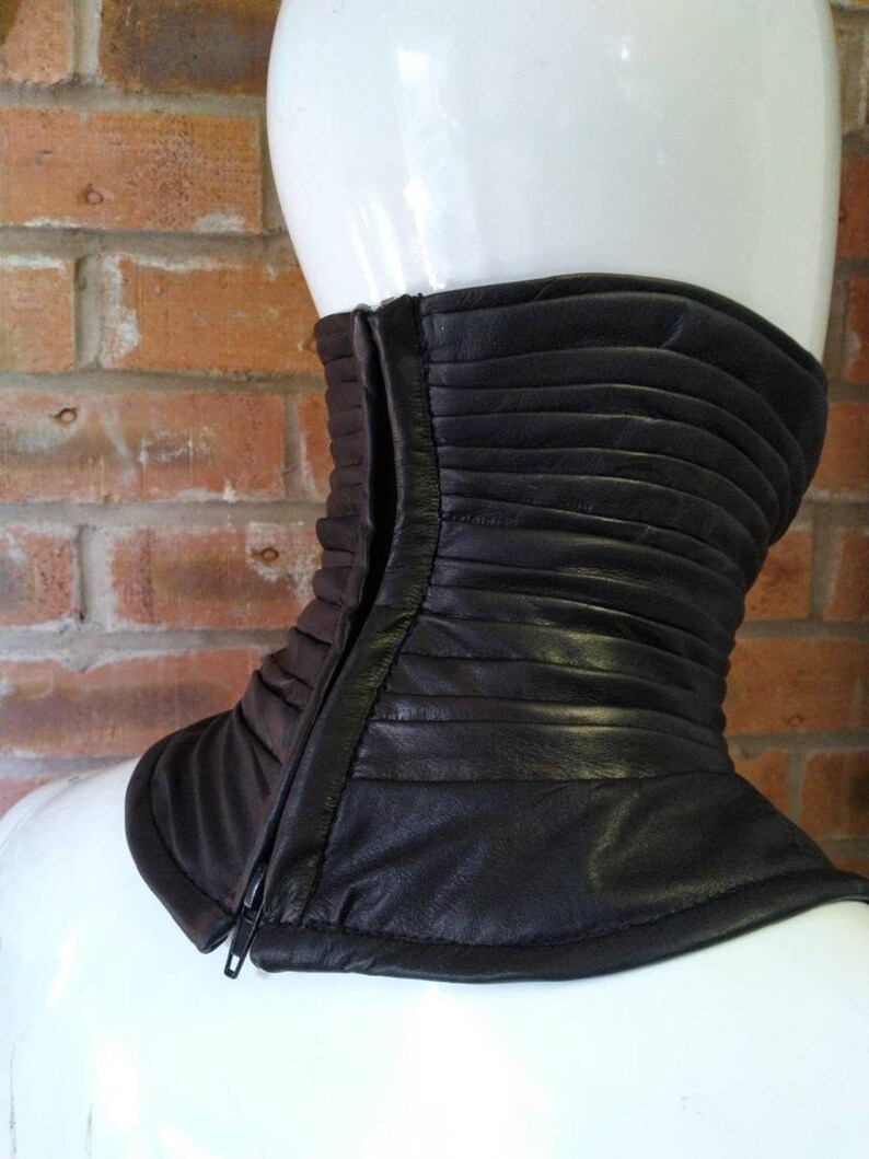 May include: A dark brown leather neck gaiter featuring horizontal pleated detailing and a vertical front zipper closure. The accessory is designed to fit snugly around the neck, providing a structured appearance with a smooth finish.